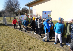 Dzieci wraz z panią maszerują z wiosennymi plakatami pod budynkiem przedszkola.